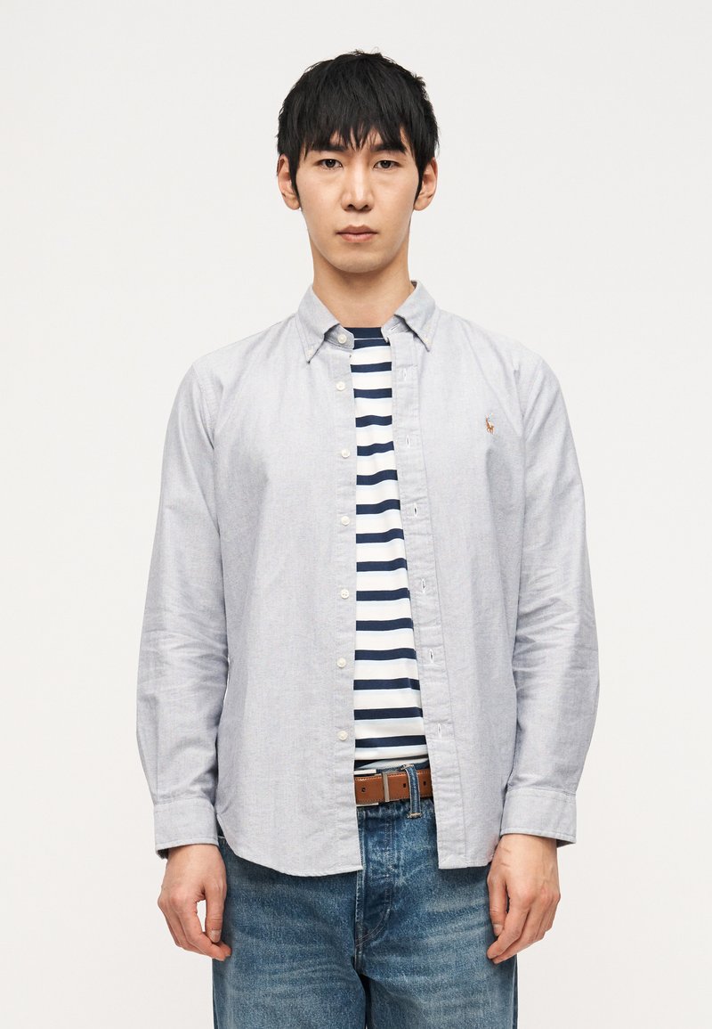 Νέος άνδρας με ανοιχτό γκρι πουκάμ ισο με κουμπιά πάνω από ένα μπλε και άσπρο ριγέ μπλουζάκι, μπλε τζιν και καφέ ζώνη, στέκεται μπροστά σε λευκό φόντο.