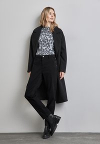 Abrigo largo negro, suéter de estampado de leopardo gris y pantalones negros con botones laterales. Modelos estilizados con botas de tobillo negras sobre fondo gris.