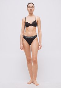 Νεαρή γυναίκα όρθια, ξυπόλητη, φορώντας μαύρο δαντελωτό σουτιέν και μαύρο εσώρουχο Polo Ralph Lauren, με λευκό απλό φόντο.