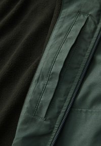 Veste vert foncé avec fermeture éclair visible et poche à fente à côté d'un tissu polaire foncé texturé, montrant des détails en gros plan des matériaux et des coutures.