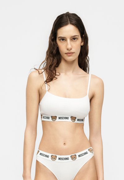 Γυναίκα που φοράει λευκό Moschino bralette και αντίστοιχα λευκά εσώρουχα με ελαστικές λωρίδες με σχέδιο αρκουδάκια και κείμενο.