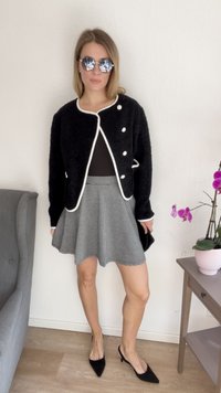 Casaco preto texturizado com acabamentos e botões brancos, combinado com uma blusa preta e uma saia plissada xadrez. Sapatos de salto preto completam o visual.