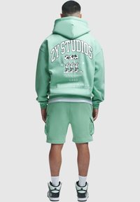 Homme portant un sweat à capuche vert menthe et un short cargo assorti, montrant le dos avec le texte « 2Y STUDIOS » et un graphisme de trois personnages tenant des globes.