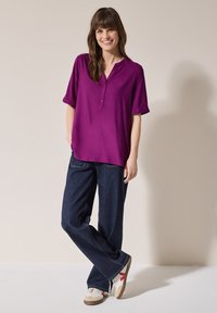 Maglietta viola a maniche corte con scollo a V e dettagli a bottoni, abbinata a jeans larghi blu scuro e sneakers multicolori.