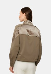 Giacca bomber marrone con parte superiore in satin lucido, polsini elasticizzati e un piccolo dettaglio metallico sul retro. Tessuto liscio e strutturato.