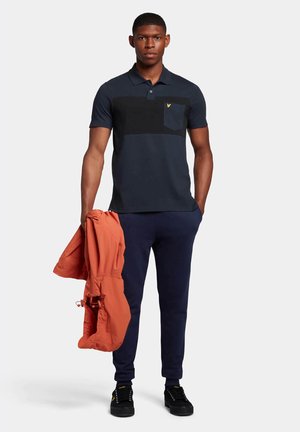Polo shirt navy con pannello orizzontale nero e tasca, abbinato a pantaloni della tuta navy. Il modello tiene in mano una giacca arancione, indossando scarpe nere.