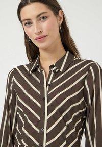 Blouse rayée marron et crème avec un devant boutonné, un col et des manches longues. Tissu lisse avec des détails en motif chevron vertical.