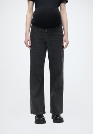 Έγκυος φορά μαύρο τζιν εγκυμοσύνης και μαύρες μπότες, στέκεται μπροστά σε ένα απλό φωτεινό φόντο.