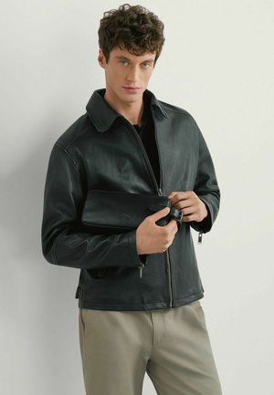 Młody mężczyzna w czarnej skórzanej kurtce i beżowych spodniach trzymający czarną skórzaną kopertówkę z klamrą na jednolitym tle.