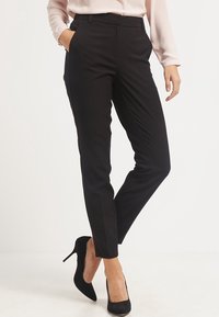 Žena v černých tailored ankle kalhotách, světle růžové blůze a černých lodičkách, stojící s jednou rukou v kapse.