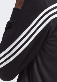 T-shirt de sport noir avec trois rayures blanches courant le long de la manche gauche, présentant une texture lisse et un design ajusté.
