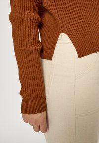 Smeđi rebrasti pulover s dugim rukavima, s razdvojenim rubom. Uparen s svjetlosmeškim hlačama visokog struka s bočnom džepom.