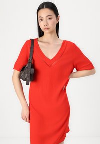 Vestido vermelho de decote em V com mangas curtas e tecido texturizado. Acentuado com costuras delicadas ao longo da gola. Mala de ombro em couro preta.