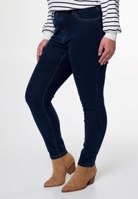 Un jean skinny bleu foncé avec une texture lisse, doté d'un design classique à cinq poches, associé à des bottines en daim beige.