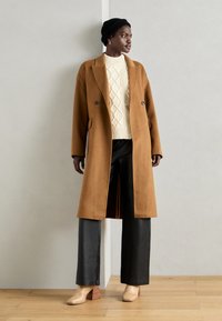 Brauner Wollmantel, cremefarbener Kabelstrickpullover, schwarze Lederhosen und hellbraune Stiefel mit Blockabsatz. Der Mantel hat ein doppelt geknöpftes Design.