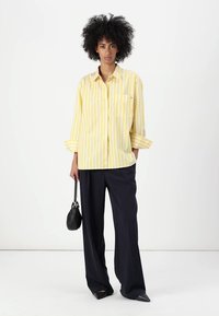 Camicia oversize a righe gialle e bianche con maniche lunghe e tasca sul petto, abbinata a pantaloni neri larghi e una piccola borsa nera.