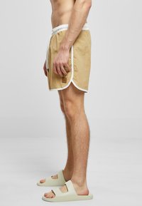 Beige badebukser med hvit kant, elastisk midje og sidelommer. Kombinert med lyse farget slidesandaler. Glatt stoff tekstur.