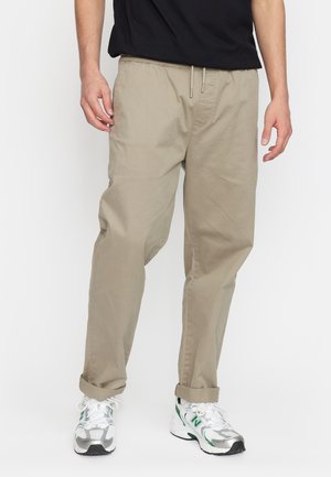 Beige, loose-fitting pants with an elastic waistband and drawstring. Rolled cuffs and a smooth fabric texture, worn with silver and green sneakers.