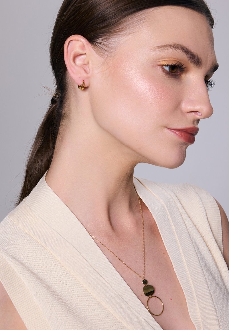 Złote kolczyki i naszyjnik z czarnym okrągłym wisiorkiem na modelce w kremowej, prążkowanej bluzce. Gładka faktura.