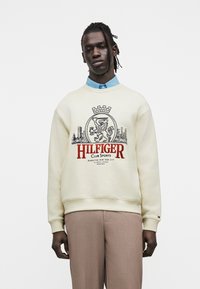 Άνδρας με πλεξούδες, φορώντας εκρού φούτερ με θυρεό λιονταριού και το κείμενο «HILFIGER Club Sports», μπλε πουκάμισο και μπεζ παντελόνι, στέκεται μπροστά σε λευκό φόντο.