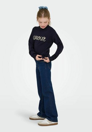 Fille ajustant un pull marine avec le texte "amour", portant un jean bleu foncé, des baskets blanches et un chouchou bleu, se tenant sur fond blanc.