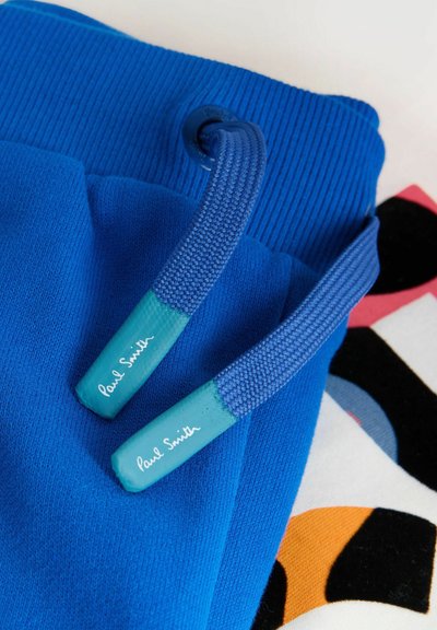 Tela azul con textura de punto que presenta cordones azules con el logo "Paul Smith" en puntas de plástico verde azulado, sobre una tela con patrón colorido.