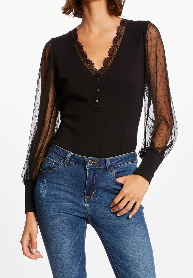 Femme portant un haut noir à manches longues avec des manches transparentes à pois et un col à volants, associé à un jean bleu, posant avec une main dans la poche.
