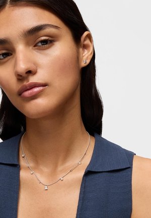 Srebrny naszyjnik z małymi okrągłymi i łezkowatymi akcentami, noszony razem z pasującymi kolczykami na sztyfcie. Modelka ma ciemne włosy, a tło jest neutralne.
