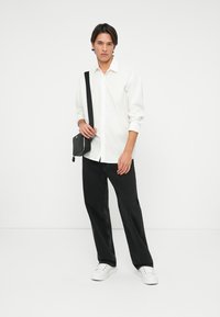 Camasa albă cu butoni, blugi negri largi și adidași albi. Geantă neagră crossbody pe umăr, cu păr drept și accesorii minime.