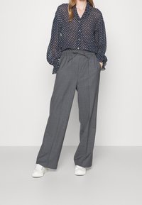 Blouse transparente bleu marine à pois avec fermeture à boutons et manches bouffantes, associée à un pantalon large taille haute gris clair et des baskets blanches.