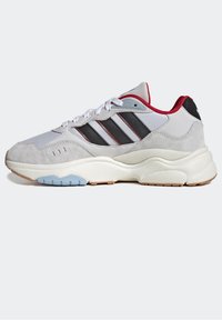 Vit och grå sneaker med röd kant, svarta ränder, vita snören och en tjock krämfärgad sula med blåa och bruna accenter.