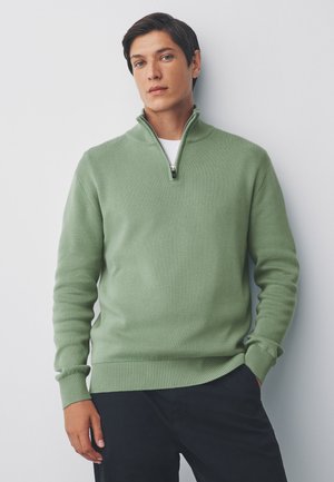 Giovane uomo che indossa un maglione verde chiaro con zip a quarto e pantaloni neri, in piedi davanti a uno sfondo bianco semplice.