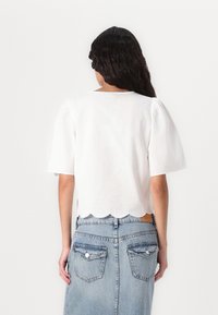 Bela bluzica s puhastimi rokavi in valovitim robom, v kombinaciji z blagim modrim jeans krilom, ki ima dva zadnja žepa in gumbasto zapiranje.