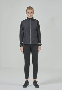 Svart lättviktsjacka med hög krage, framdragkedja och reflekterande vita ränder. Kombinerad med svarta leggings och sportskor.