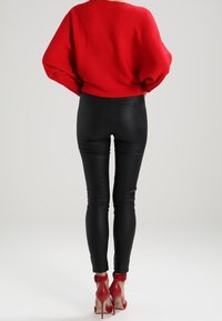 Rode oversized gebreide trui gecombineerd met zwarte leren skinny broeken en rode hoge hakken met enkelbandjes. Eenvoudige achtergrond.