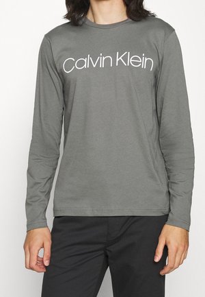 Personne portant un T-shirt gris à manches longues avec le logo blanc "Calvin Klein" sur la poitrine et un pantalon noir, debout immobile.