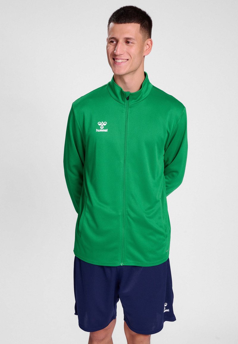 Veste zippée verte en tissu lisse, dotée d'un col haut et d'un logo blanc. Associée à un short marine. Design simple et sportif.