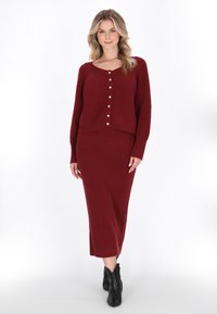 Burgundy stickad cardigan med rund halsringning och knäppning, i kombination med en matchande ribbad kjol. Modellen har på sig svarta ankle boots.