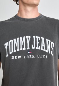 Gray cotton t-shirt featuring bold white "TOMMY JEANS" logo and "NEW YORK CITY" text. Round neckline and relaxed fit. Subtle texture detail.