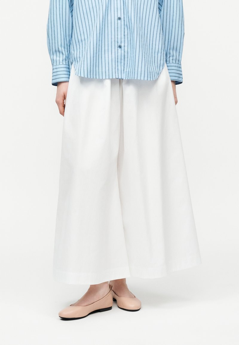 Γυναίκα που φοράει μπλε ριγέ πουκάμισο με κουμπιά, λευκό παντελόνι φαρδύ, και γυναικεία Nude παπούτσια, στέκεται σε λευκό φόντο.