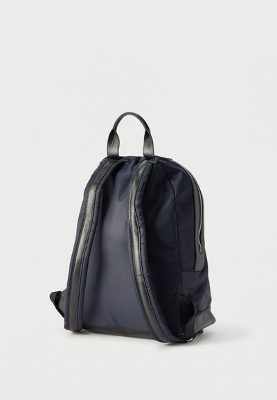 Σκούρο μπλε σακίδιο με επενδυμένους μαύρους ιμάντες, χειρολαβή στο επάνω μέρος και εμφανές φερμουάρ στο πλάι, όρθιο σε λευκό φόντο.