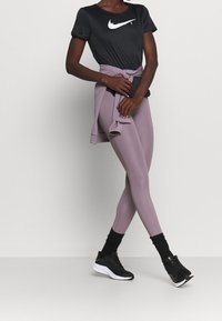 Person som bär en svart Nike-t-shirt, mauvafärgade leggings, en matchande sweatshirt knuten i midjan, svarta strumpor och svarta Nike-sneakers, stående mot en neutral bakgrund.