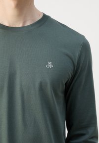 Langærmet, mørkegrøn t-shirt lavet af glat bomuld, med et lille hvidt logo "MOP" på venstre bryst. Rund halsudskæring, tætsiddende pasform.