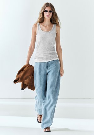 Giovane donna con occhiali da sole, canotta grigia, jeans blu larghi, giacca marrone in mano e sandali che cammina su pavimento bianco.