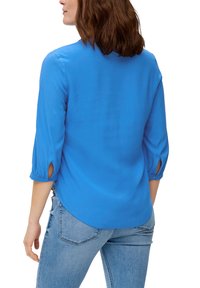 Blauwe blouse met driekwart mouwen, geknoopte manchet en een gebogen zoom. De stof lijkt soepel en lichtgewicht. Gecombineerd met lichtblauwe jeans.