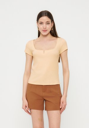 Ung kvinde med langt, glat hår iført en beige kortærmet top med firkantet halsudskæring og brune skræddersyede shorts, stående mod en hvid baggrund.