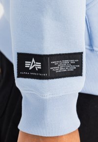 Világoskék pulóver, ribbelt mandzsettával. Fekete címkével rendelkezik, fehér szöveggel és logóval, amely a márkát és a részleteket jelzi. Puha, texturált anyag.