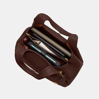 Hnedá kožená kabelka s krátkymi rúčkami, obsahujúca smartfón, pero a niekoľko zvinutých a plochých predmetov vo vnútri.