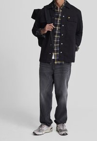 Chaqueta negra con broches plateados, camisa a cuadros en amarillo y azul marino, vaqueros grises y zapatillas grises con detalles, complementada con una mochila oscura.