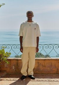 Lys beige oversized t-skjorte med en solgrafikk, kombinert med løse kremfargede bukser og svarte slip-on-sko, satt mot en kystbakgrunn.